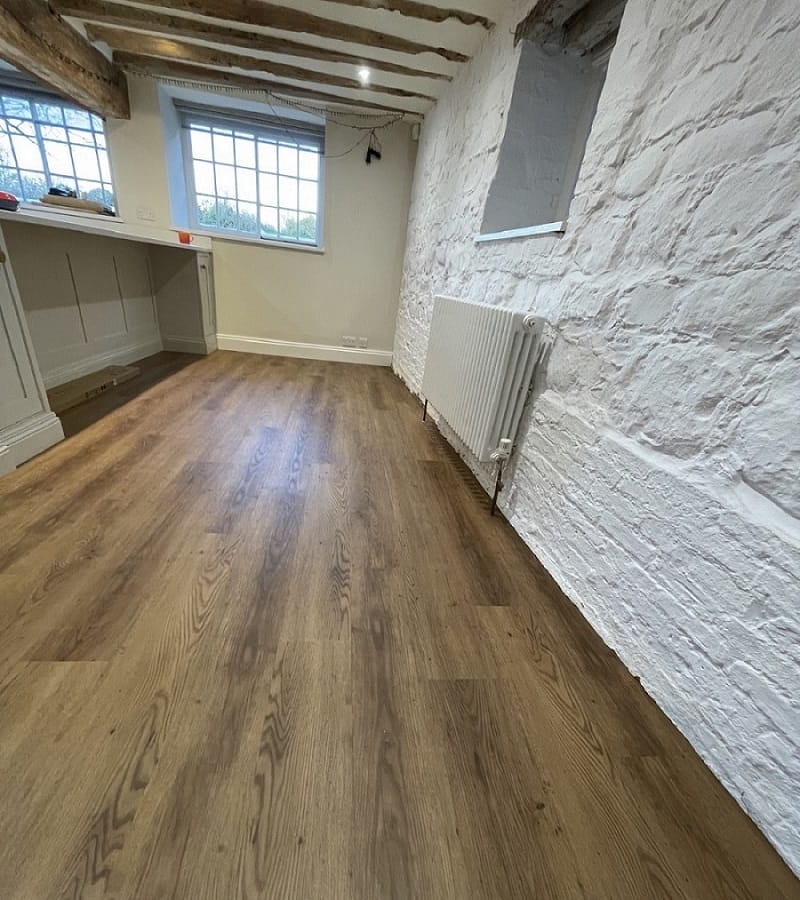 Low-angle view of Croft Oak LVT planks running the length of a Cambridge kitchen beside a white stone wall and radiator