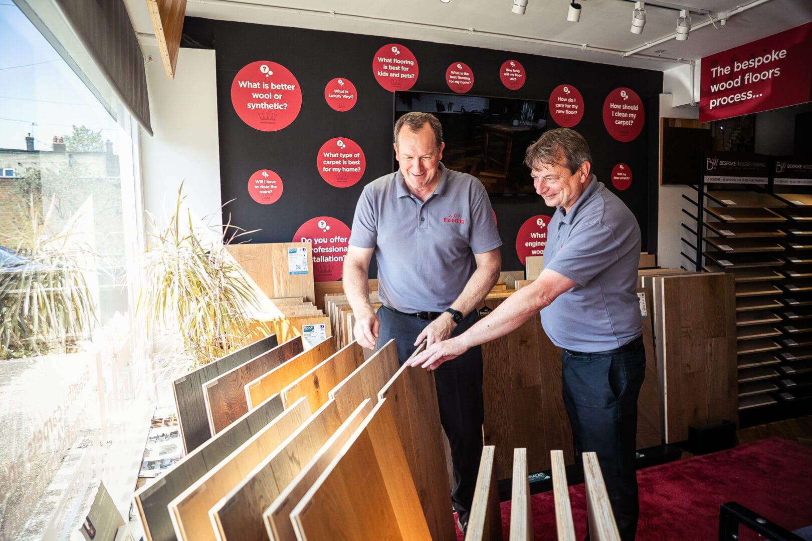 Two Art of Flooring team members examining wood flooring samples together in the showroom, with FAQ signage on the wall behind them