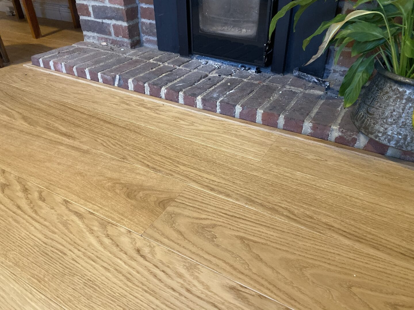 Light oak wood flooring meeting a brick fireplace hearth, with a wood-burning stove and a large leafy plant nearby
