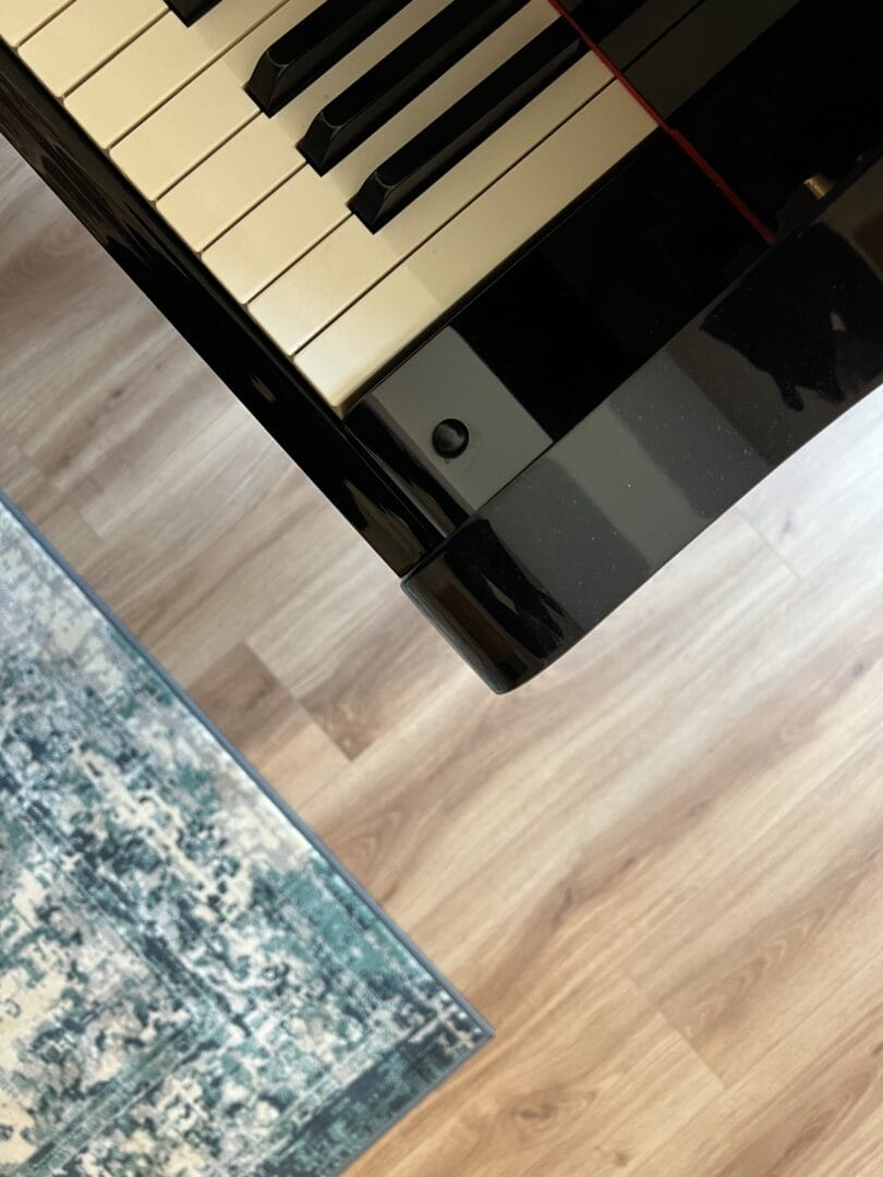 Close-up of a grand piano's keys and black casing resting on light oak wood flooring, with the edge of a teal rug nearby