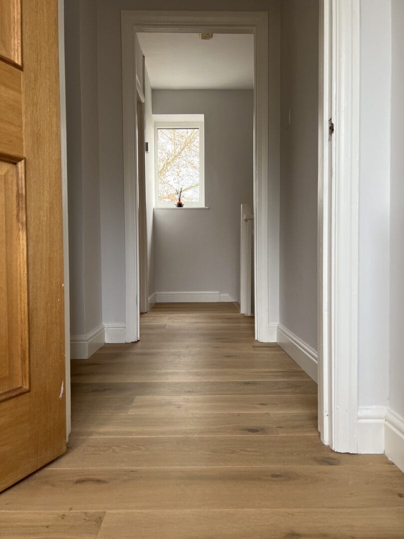 A bright upstairs hallway with light oak wood flooring, white painted doors, and a small window at the far end