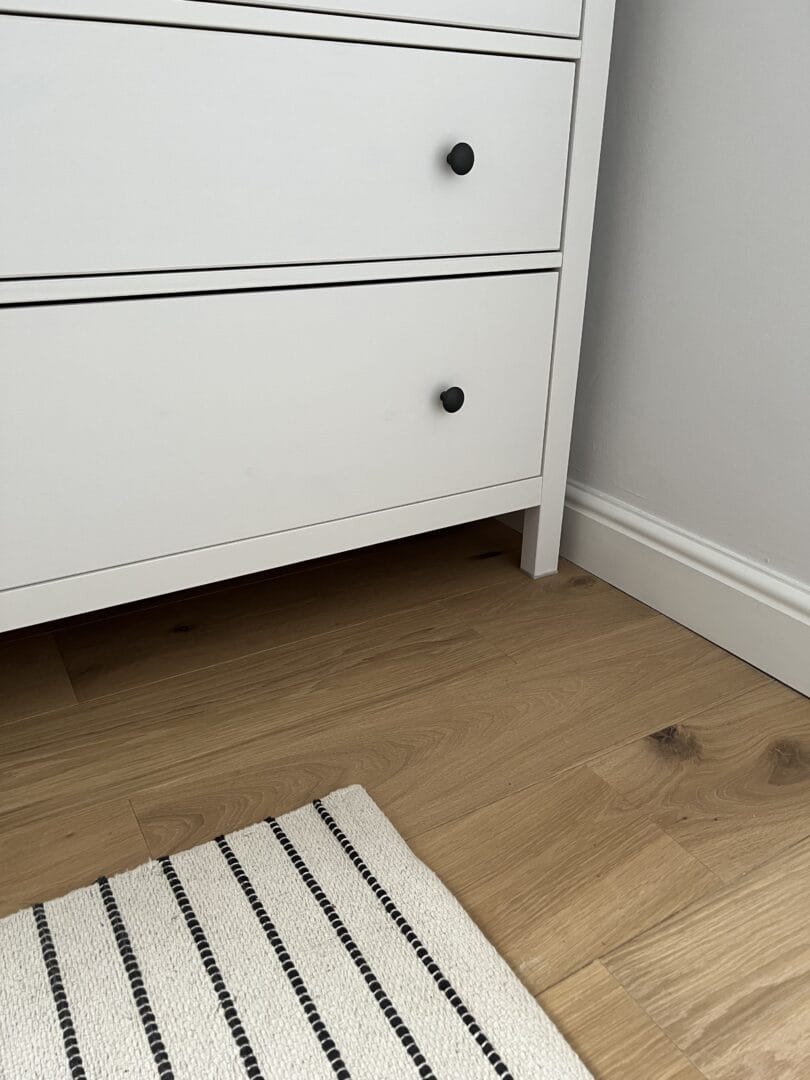 Corner of a white chest of drawers with black knobs standing on light oak wood flooring, next to a black and white striped rug