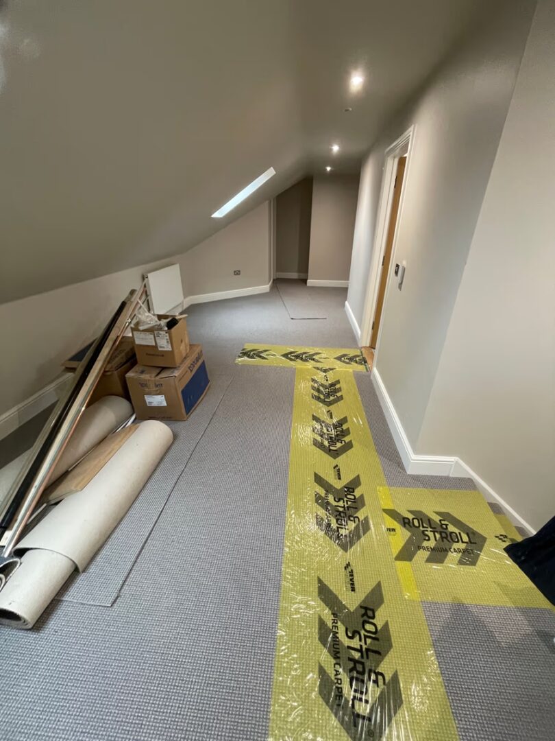 A long landing corridor with newly fitted grey carpet, protected by a strip of film, with boxes and building materials stacked to one side.