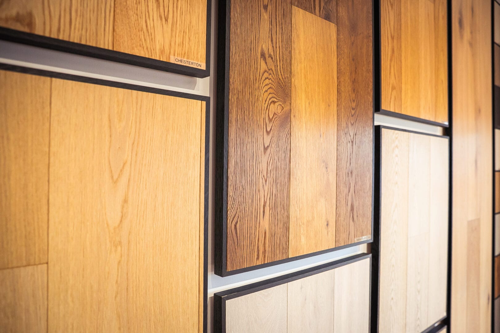 Framed oak flooring samples on a showroom wall, including a panel labelled Chesterton, in honey, mid-brown and pale finishes.