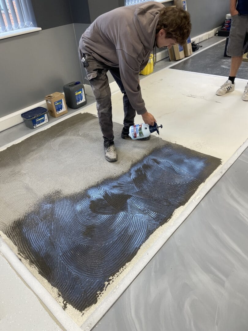 Young fitter using a hand pump sprayer over a freshly combed floor surface marked with curved trowel lines.
