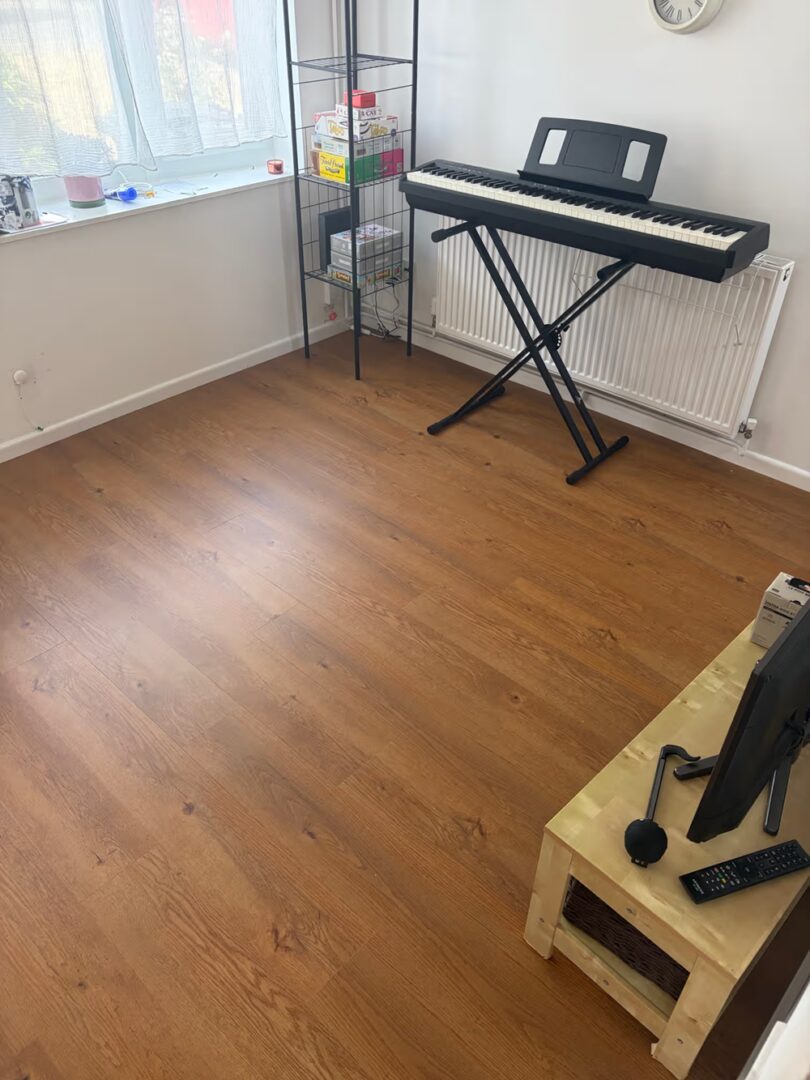 Bright living room with oak effect vinyl planks, a digital piano by the radiator and a window with sheer curtains.