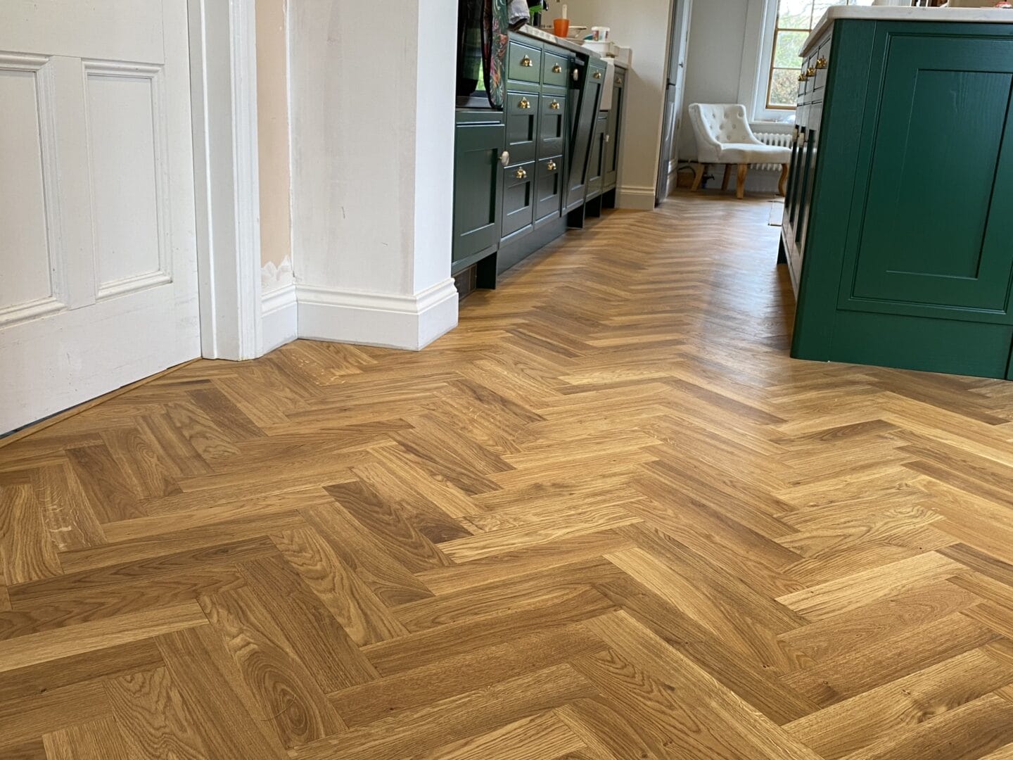 Oak herringbone flooring running through a kitchen with dark green shaker-style cabinets and a matching green island.