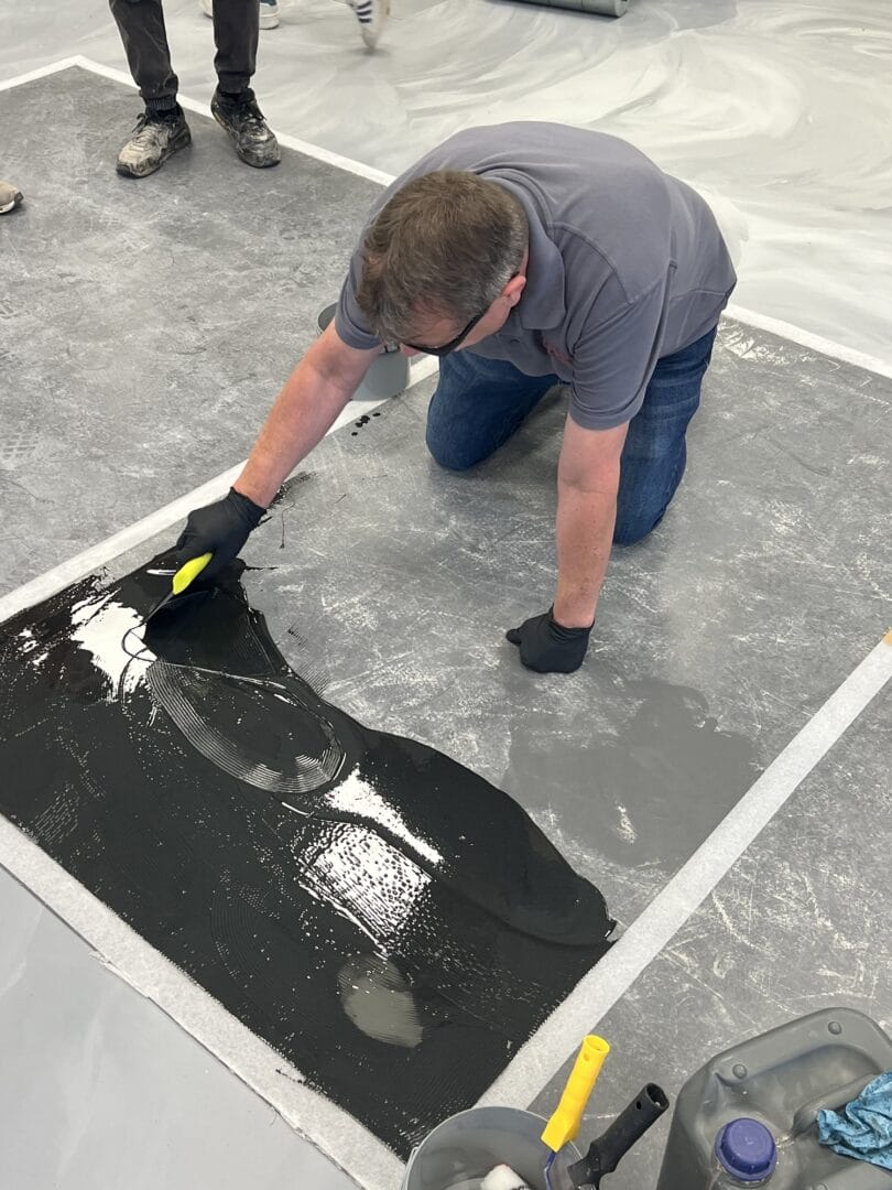 Fitter spreading a glossy black coating across a taped-off section of floor with a notched trowel.