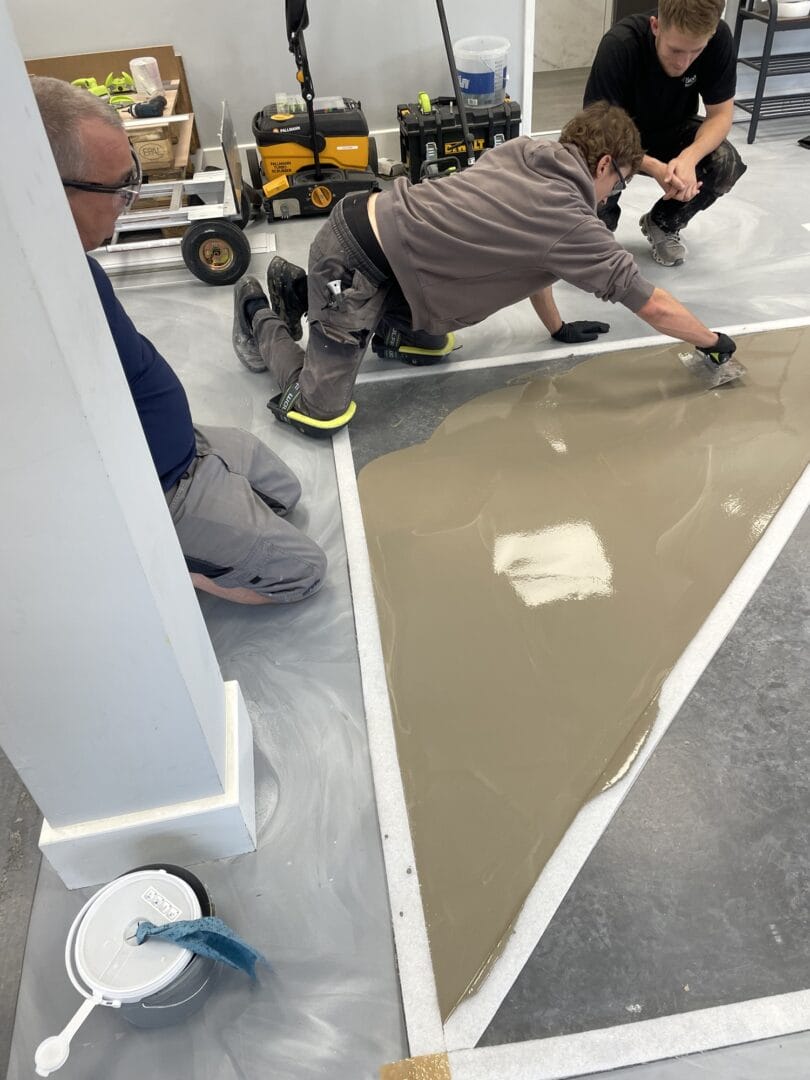 Trainee in knee pads spreading levelling compound with a trowel across a triangular taped-off section while two others watch.