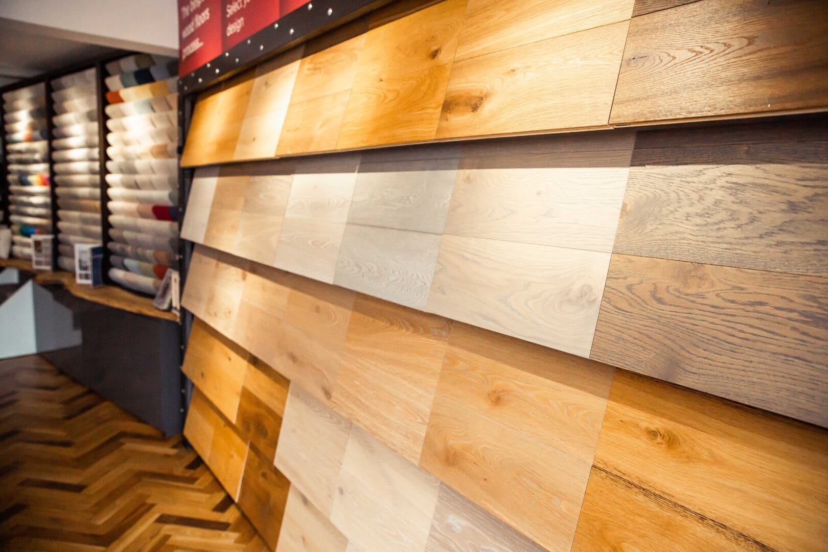 Wall of oak flooring samples in light, natural and grey finishes, with carpet sample racks behind and a herringbone floor below.